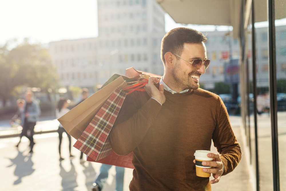 A smiling man wearing sunglasses and carrying multiple shopping bags over his shoulder while holding a takeaway coffee cup outside a store window.
