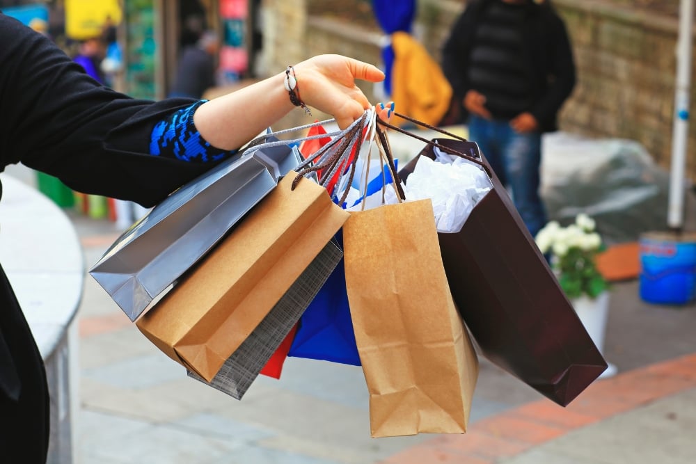 Close-up of a person's hand holding a bundle of colourful paper shopping bags outdoors.