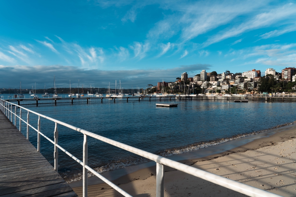 A wooden pier with white railings along the sandy shoreline of Redleaf Beach, with a netted harbour pool and hillside apartments in the background.