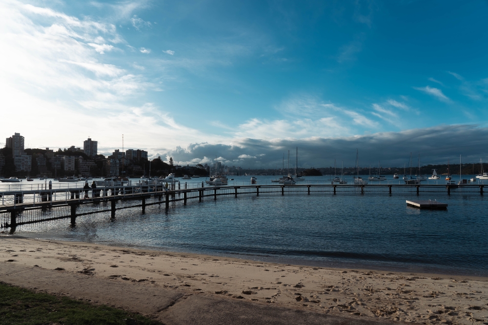 A curved wooden boardwalk and shark net enclosure extending from a sandy beach into a calm bay with anchored sailboats.