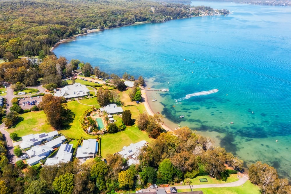 Aerial view of clear turquoise water along a sandy bay, bordered by lush green lawns, coastal homes, and dense forest.